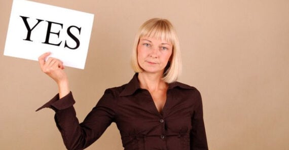 A woman with blond hair wearing a brown shirt holds up a white sign with the word "YES" in bold black letters. She stands against a plain beige background and has a neutral expression.
