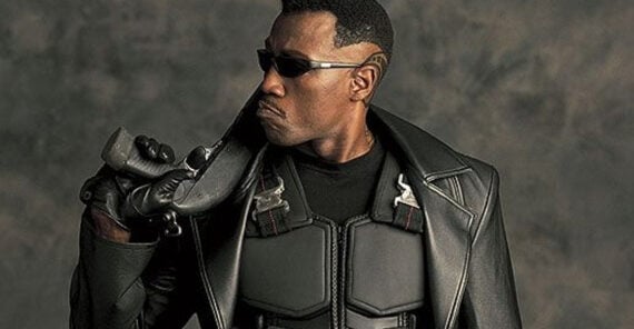 A man wearing black sunglasses, a black leather coat, and body armor holds a sword over his shoulder. He has a serious expression and a distinct haircut with patterns shaved into the sides. The background is gray and textured.