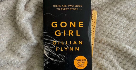 A copy of the novel "Gone Girl" by Gillian Flynn rests on a textured, light-colored blanket. The book cover is black with bold yellow text and a round sticker stating "Thriller of the Year.