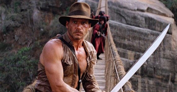 A man in a brown hat and rugged shirt stands on a rope bridge holding a large machete, with cliffs in the background and several people dressed in red approaching from behind.
