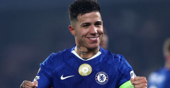 A soccer player wearing a blue Chelsea FC jersey smiles and raises his fists in celebration. He has short dark hair, visible tattoos on his neck, and a FIFA gold badge on his chest.