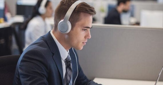 A man in a suit wearing headphones sits at a desk in an office cubicle, focused on his computer. Other people are visible working in the background.