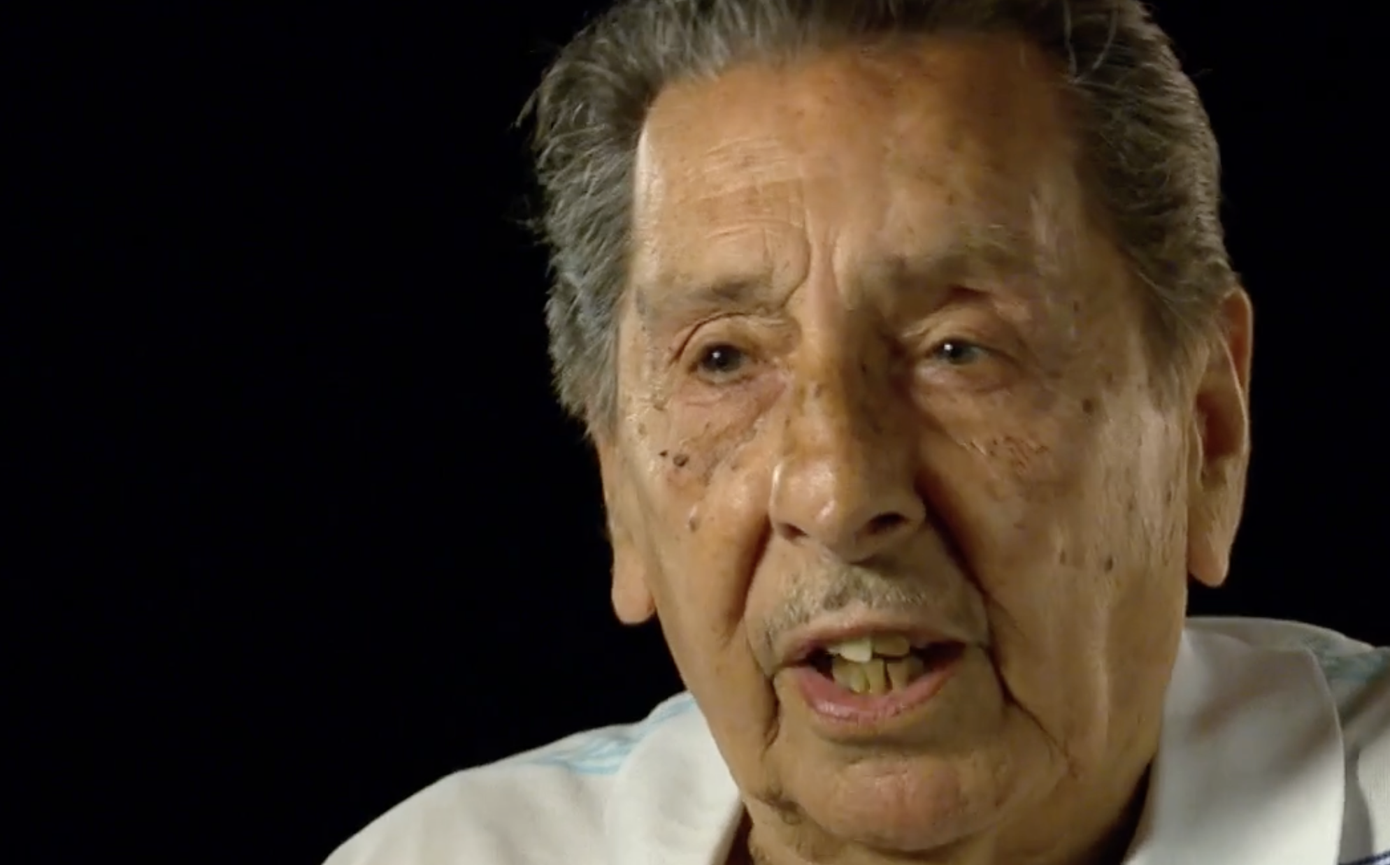 An elderly man with gray hair and a mustache is speaking. He is wearing a light-colored shirt and is set against a dark background. His expression appears serious and engaged.