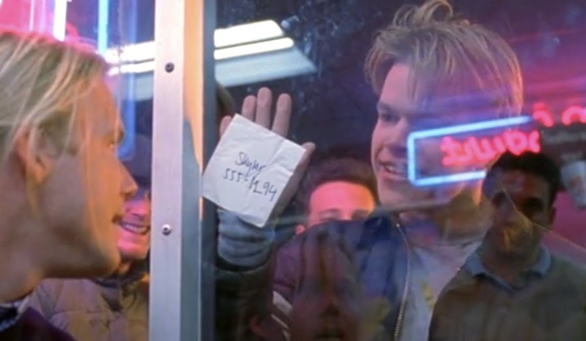A young man smiles and presses a napkin with a phone number written on it against a glass window while talking to someone outside. Neon lights glow in the background.