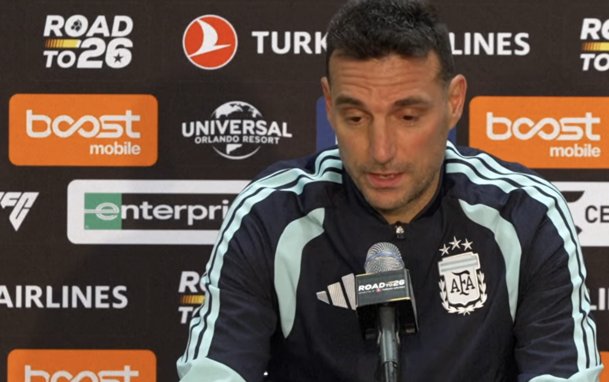 A man in an Argentina football jacket speaks at a press conference, sitting in front of microphones and a backdrop with various sponsor logos.