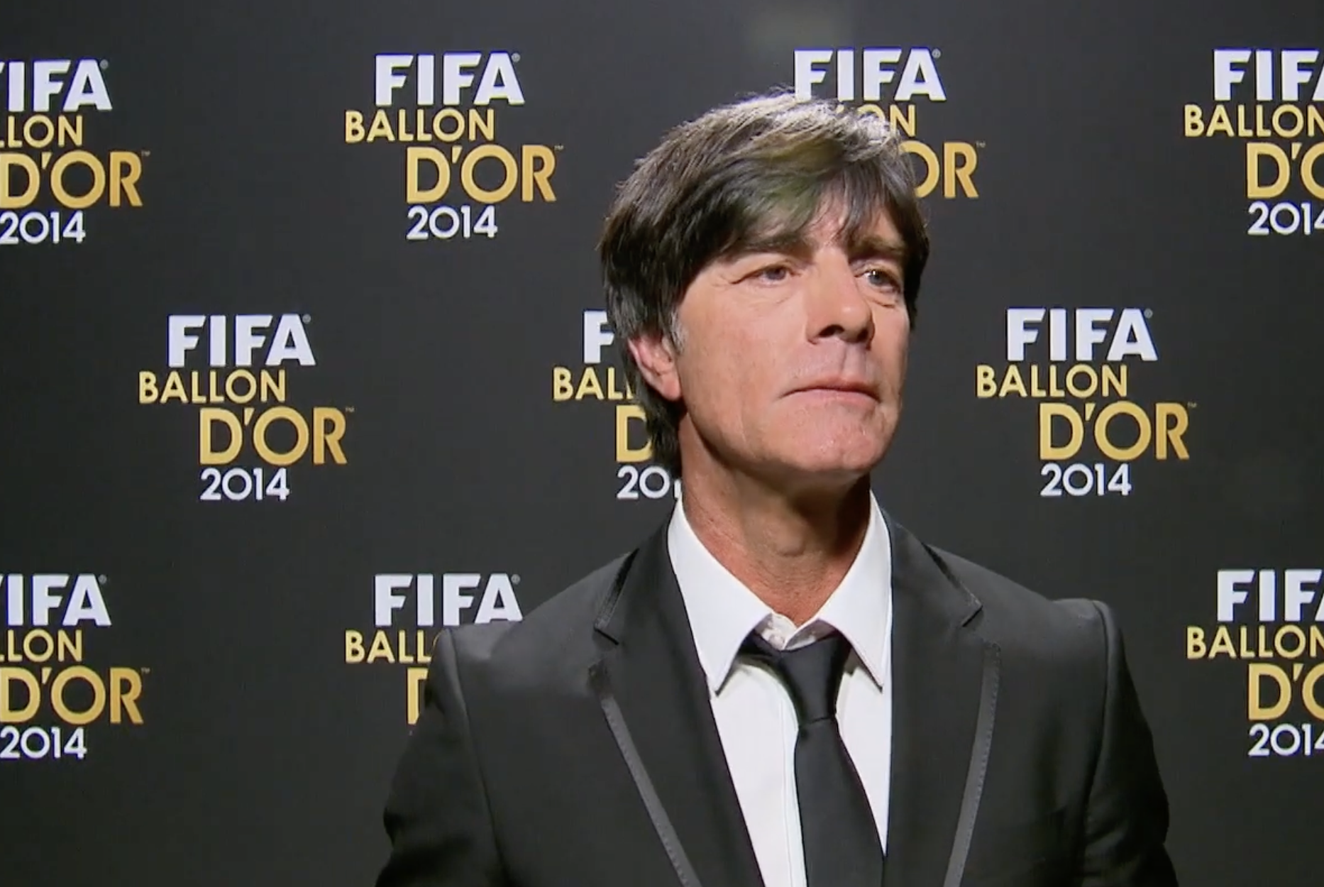 A man in a black suit and tie stands in front of a backdrop with the text "FIFA Ballon d'Or 2014" repeatedly printed in gold and white.