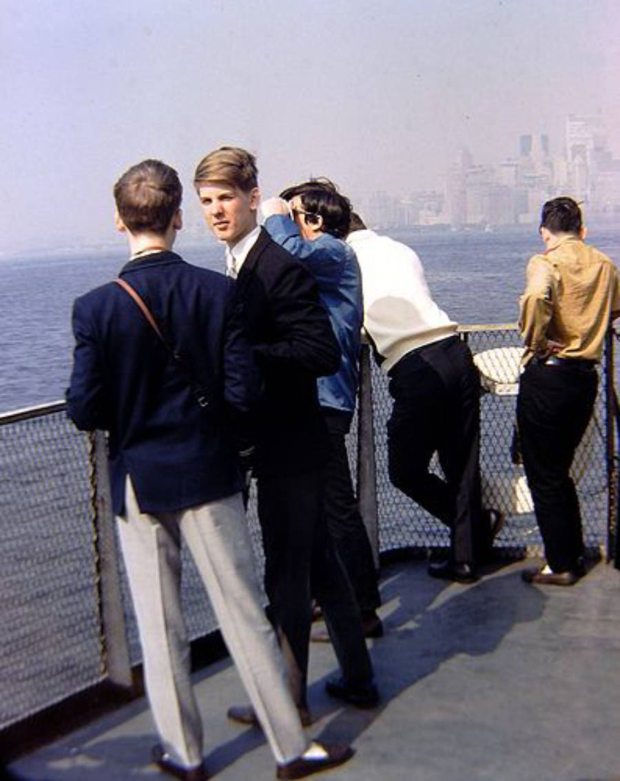 Four young men stand on the deck of a ferry, some looking out over the water while others talk. A hazy city skyline is visible in the background. The scene suggests a casual day by the waterfront.
