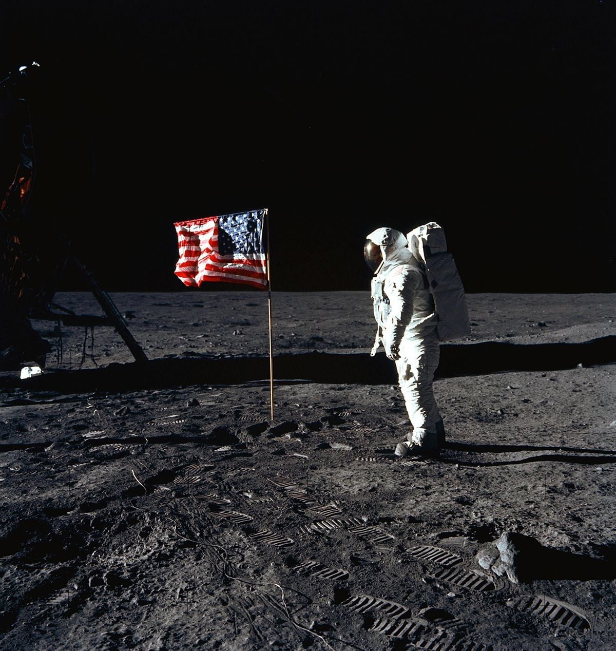 An astronaut in a spacesuit stands on the Moon next to an American flag, with lunar lander parts and footprints visible on the gray, dusty surface against a black sky.