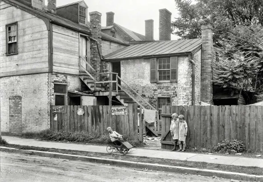 Two young girls stand by a wooden fence in front of old, weathered brick houses. A child in a stroller is pushed along the sidewalk. The area appears run-down, with peeling paint, exposed bricks, and wooden stairs.