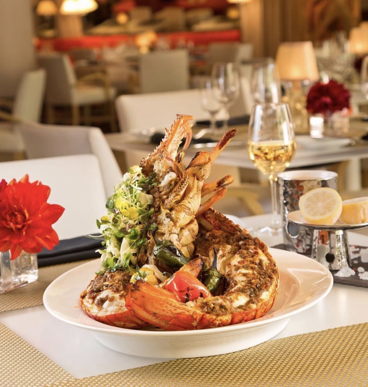 A gourmet lobster dish garnished with greens and vegetables sits on a white plate at an elegant restaurant table, accompanied by a glass of white wine, candles, and red flower decor.