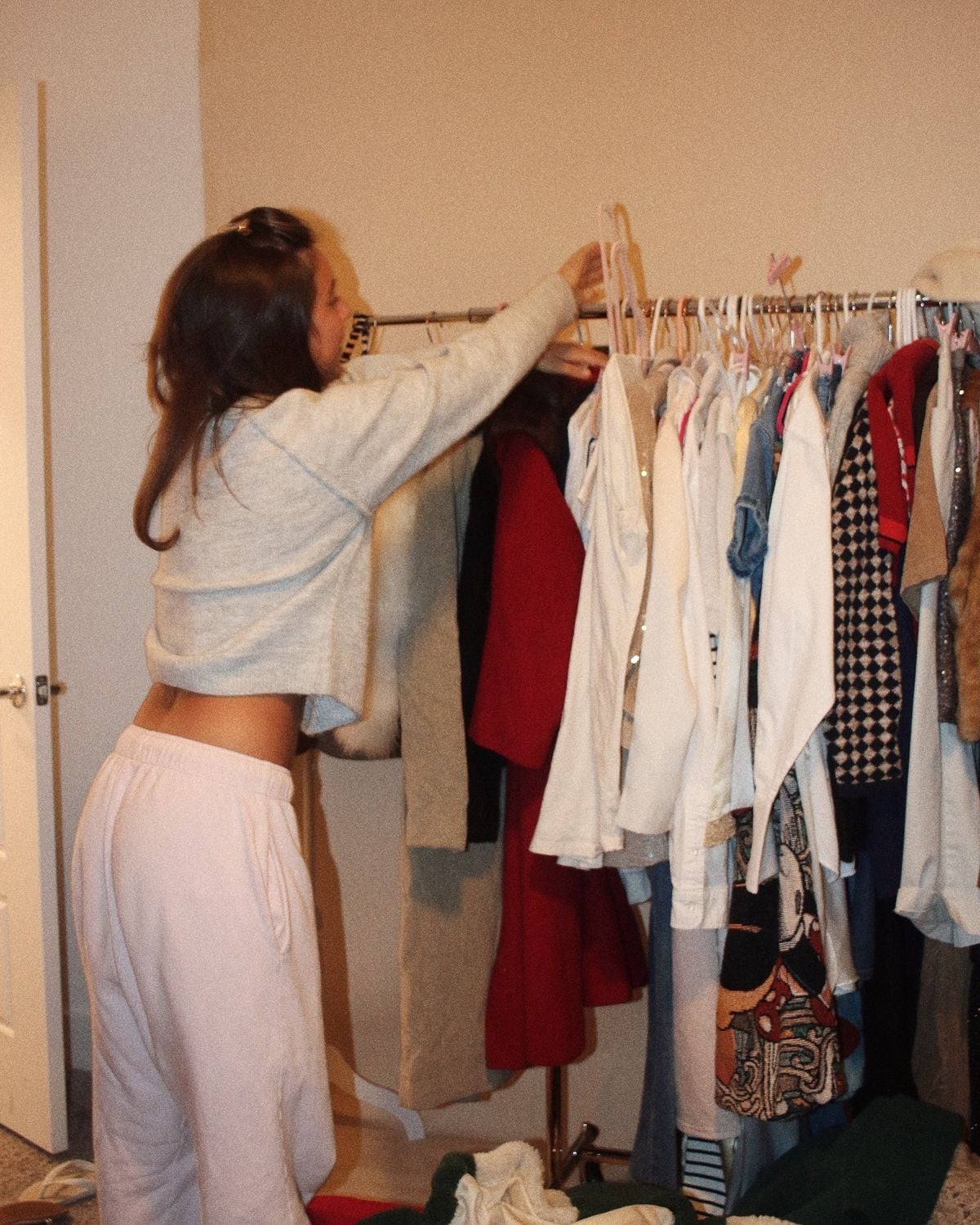 A person in a gray cropped sweater and light pants is reaching to hang clothes on a rack filled with various garments in a room with beige walls.