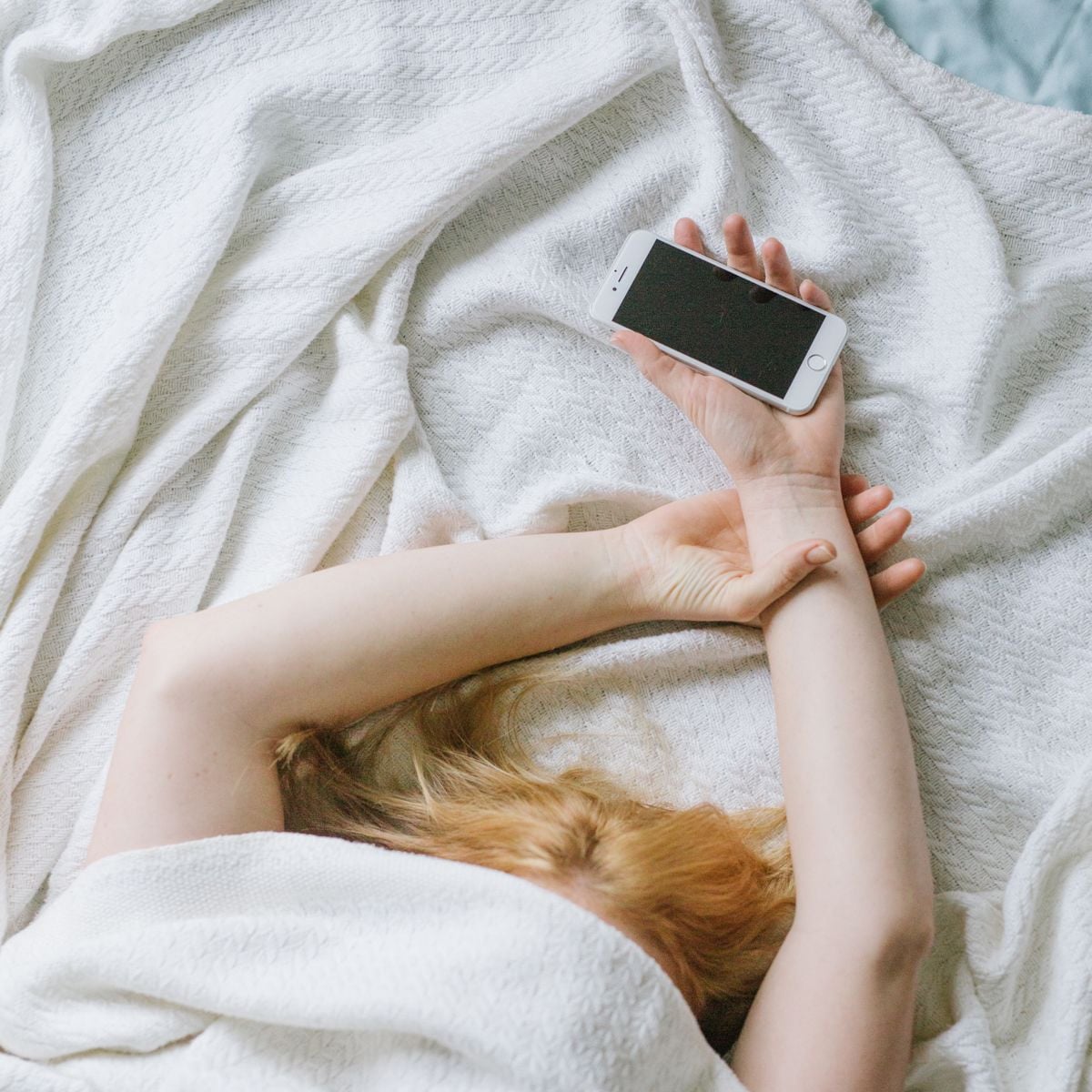 A person with long light hair lies in bed under a white blanket, holding a smartphone in one hand above their head. The phone screen is off, and the person appears relaxed or resting.