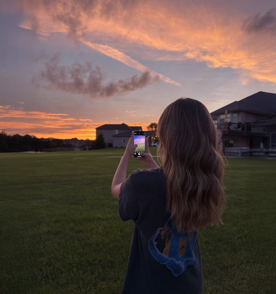 A person with long, wavy hair stands on a grassy lawn at sunset, taking a photo of the colorful sky with their phone. Houses are visible in the background under the vibrant orange and purple clouds.