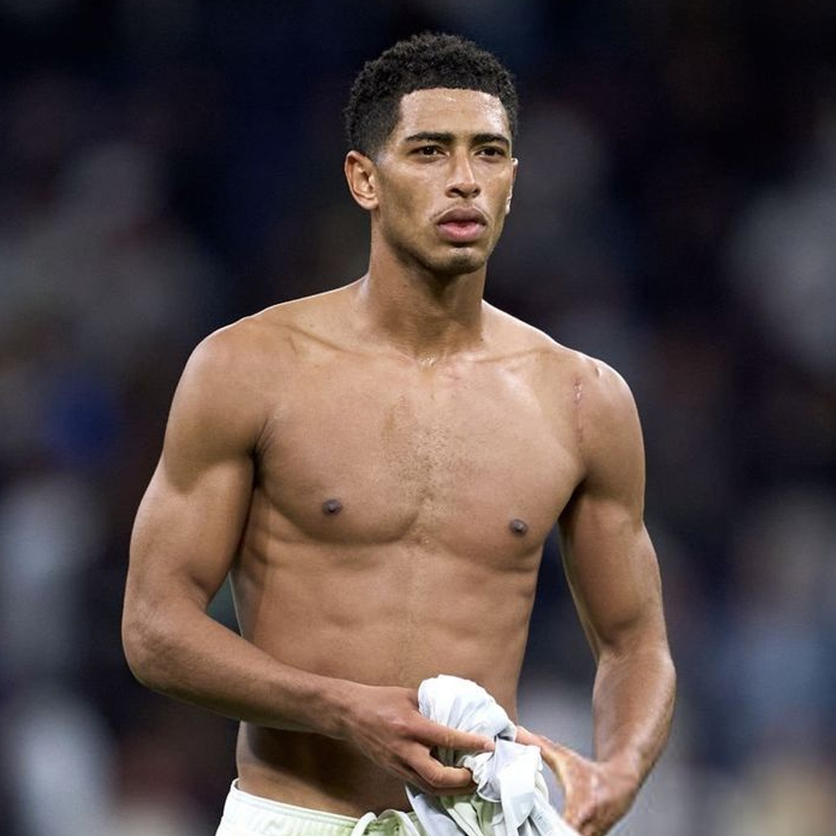 A shirtless male athlete with short curly hair stands on a sports field, holding a white shirt in his hand, looking focused and slightly tired. The background is blurred with indistinct spectators.