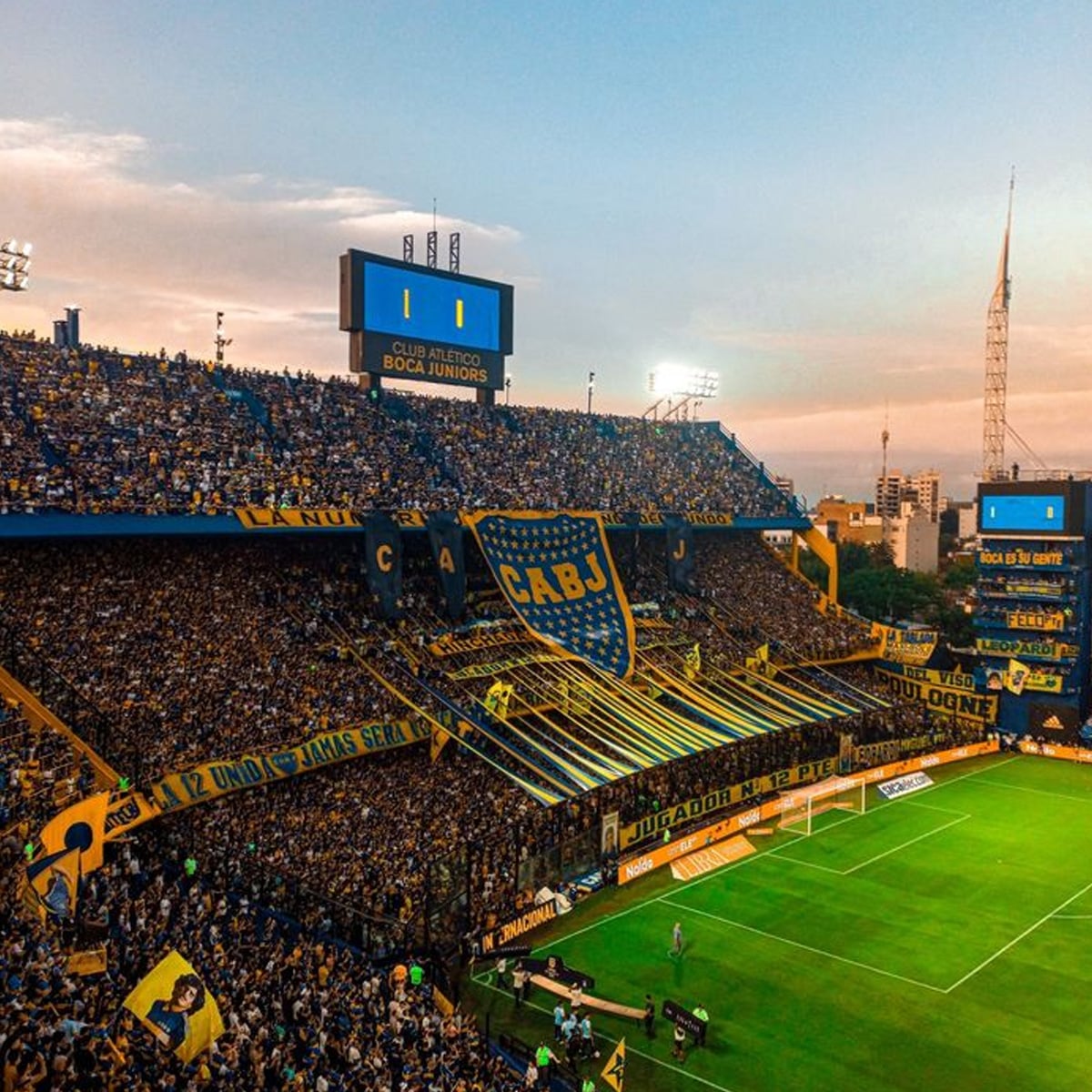A packed soccer stadium filled with Boca Juniors fans waving flags and banners, with blue and yellow colors dominating the stands. The scoreboard shows 0-0 and the atmosphere is lively at sunset.