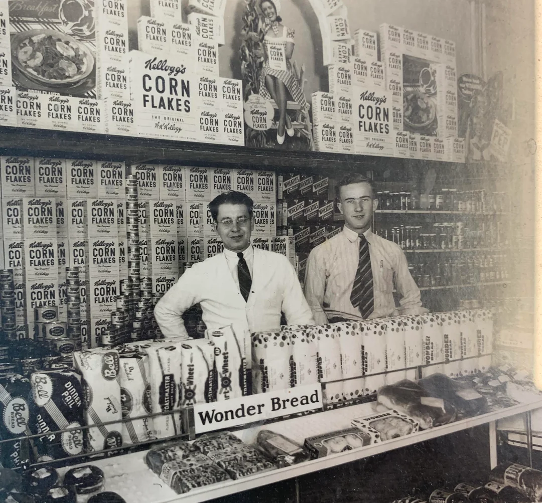 Two men in dress shirts and ties stand behind a grocery display filled with Wonder Bread loaves and pastries. The shelves and wall behind them are stacked high with Kellogg’s Corn Flakes cereal boxes.