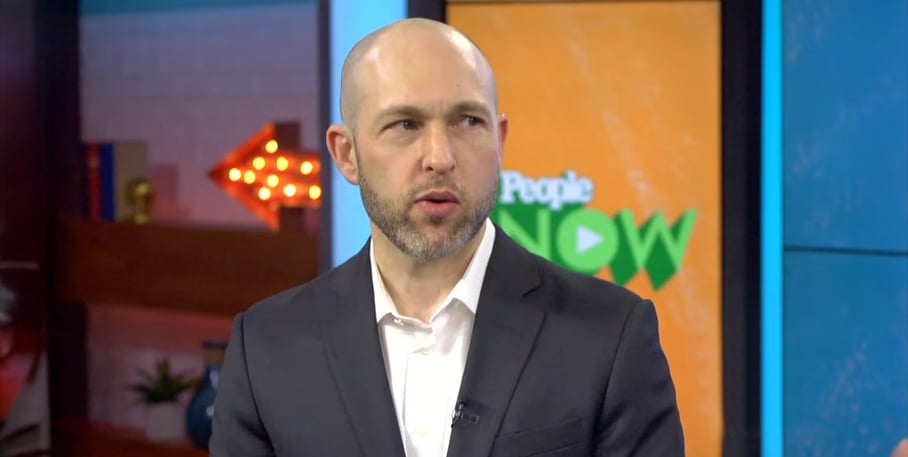 A man in a dark suit and white shirt speaks in a studio with colorful set decor. A screen behind him displays the "People NOW" logo, and a lit arrow sign is visible in the background.