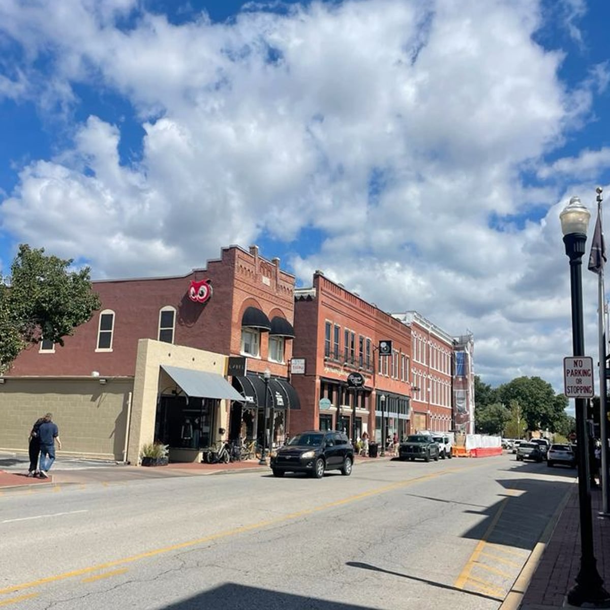A sunny street scene in a small town shows brick storefronts, parked and moving cars, a few pedestrians, and a sky filled with fluffy clouds. A "No Parking" sign is visible on the right side of the image.
