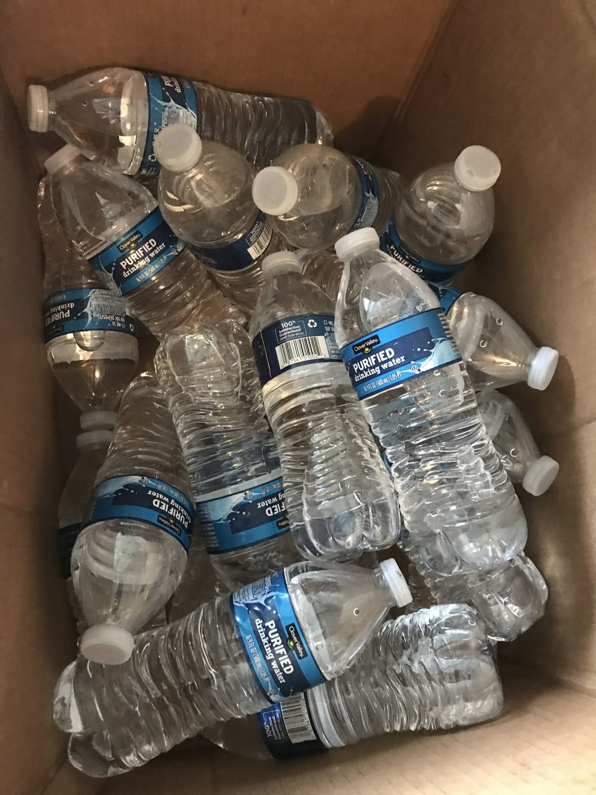 A cardboard box filled with several clear plastic water bottles, some standing upright and others lying on their sides. The bottles have blue labels and white caps.