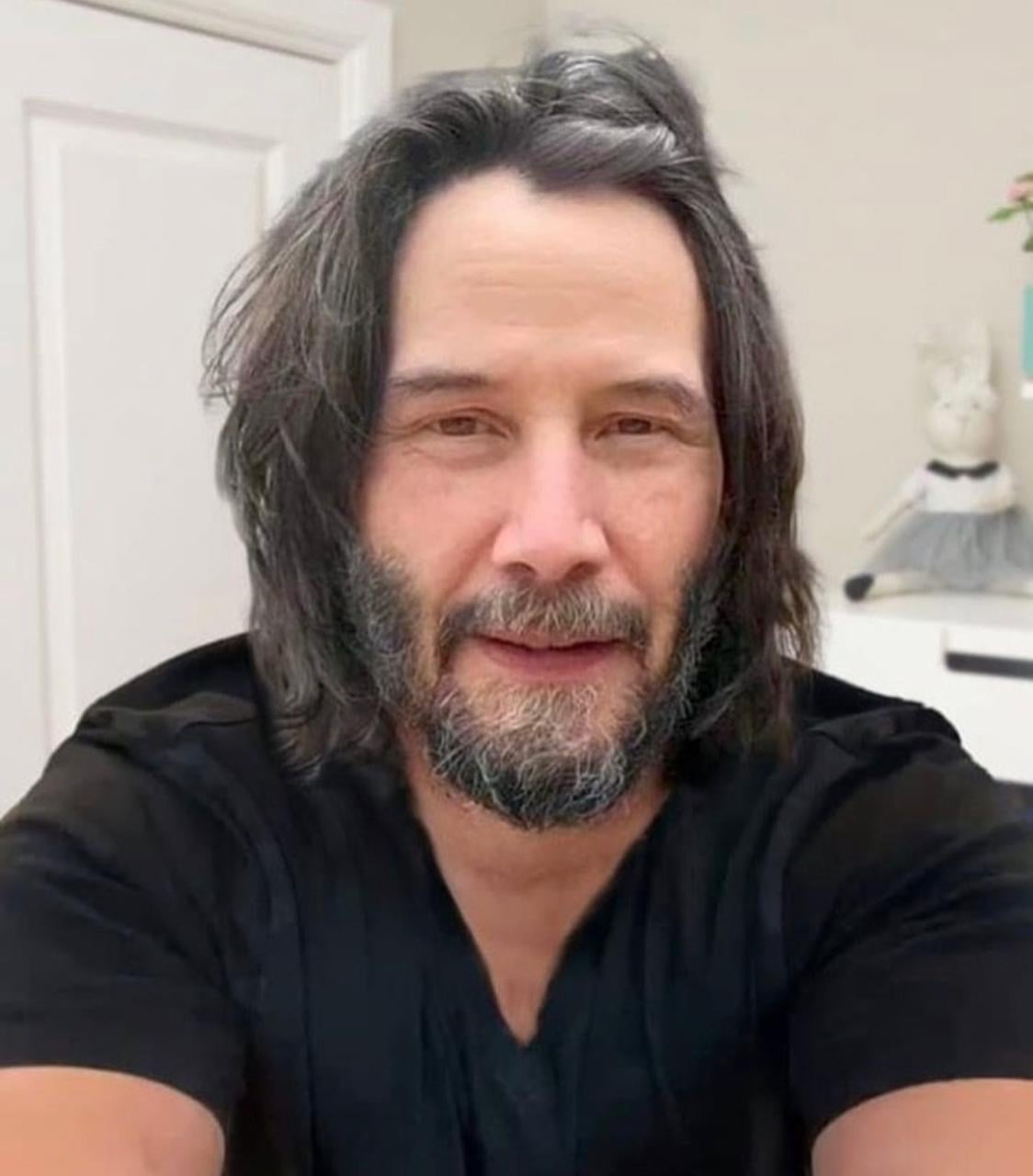 A man with shoulder-length dark hair and a beard is wearing a black shirt, sitting indoors and smiling slightly at the camera. A stuffed toy in a dress is visible on a white cabinet in the background.