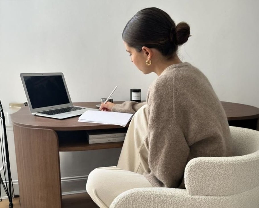 A woman with dark hair in a bun sits at a desk, writing in a notebook with a pen. An open laptop is on the desk, and she is facing away from the camera. The setting appears to be a home office.