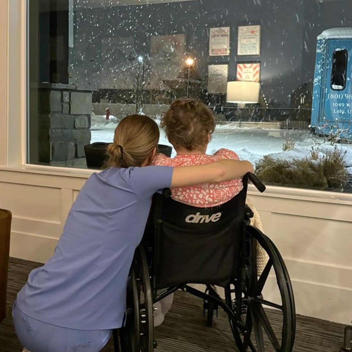 A caregiver in scrubs hugs an elderly woman in a wheelchair as they look out a large window at snowfall and a snowy scene outside at night.