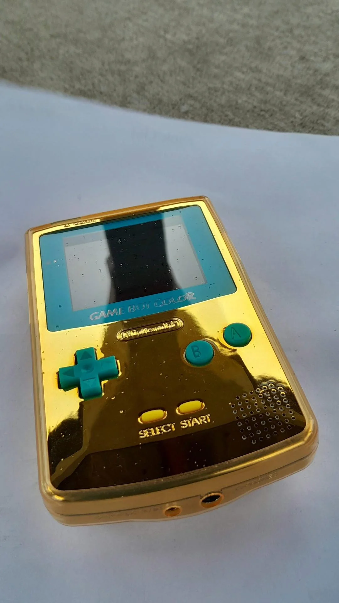 A shiny gold Game Boy Color handheld console with turquoise buttons rests on a white surface, viewed from an angle.