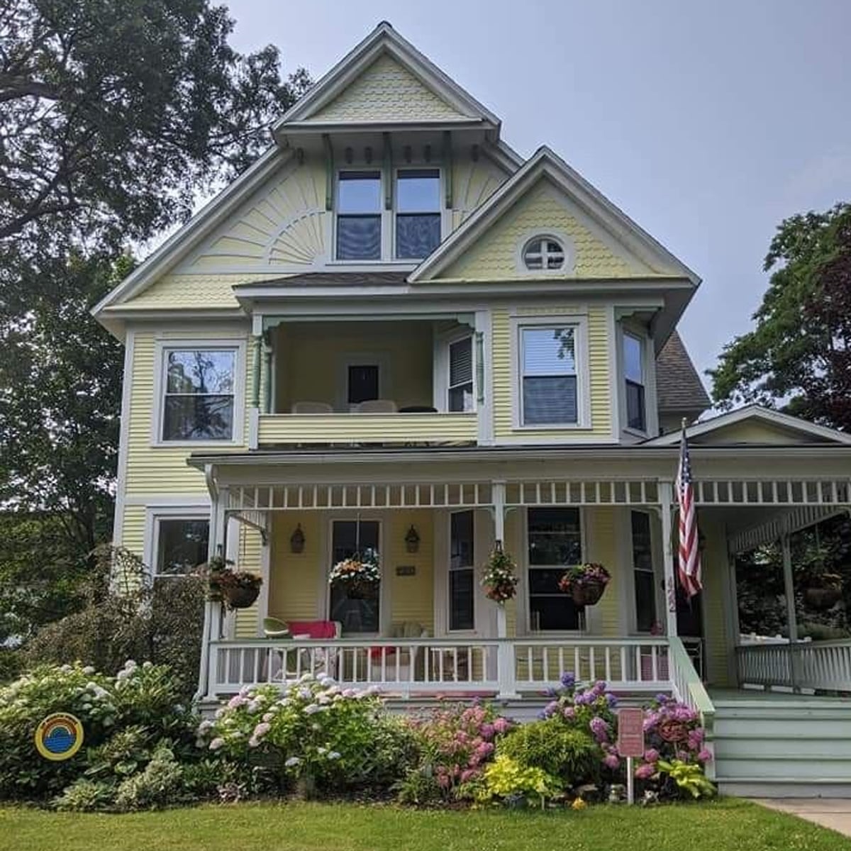 A large yellow Victorian-style house with white trim, a wraparound porch, potted flowers, a green lawn, and an American flag. There are two stories plus an attic and various blooming plants in front.