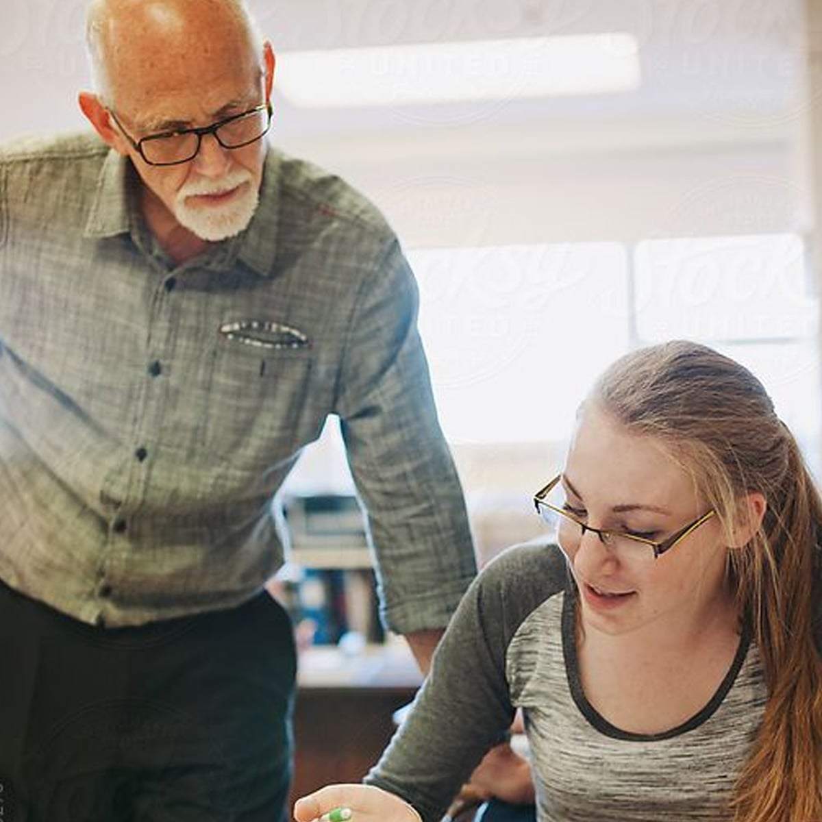 An older man with glasses leans over to help a young woman with long hair and glasses, who is seated and holding a pen, in a well-lit room. They appear to be discussing something on her paper.