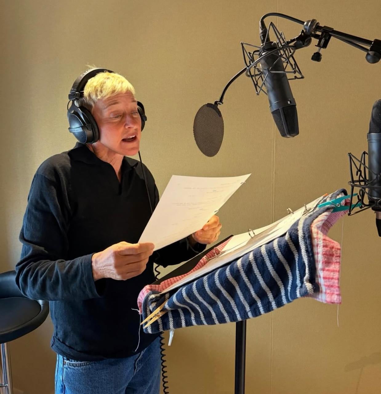 A person with short gray hair wearing headphones stands at a microphone, reading from a script on a music stand covered with striped cloths in a recording studio.