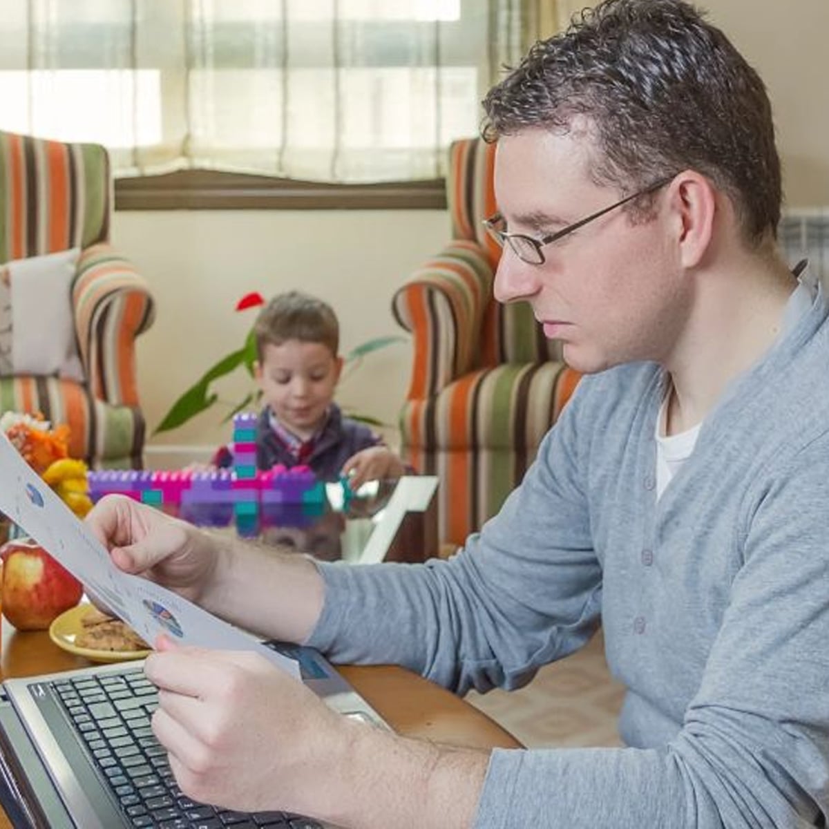 A man wearing glasses works at a laptop and looks at papers, while a young boy plays with colorful blocks in the background in a living room.