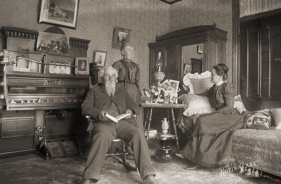 A sepia-toned photo shows an older man sitting in a rocking chair with a book, while two women in Victorian clothing stand and sit nearby in a well-decorated parlor with ornate furniture, framed pictures, and a piano.