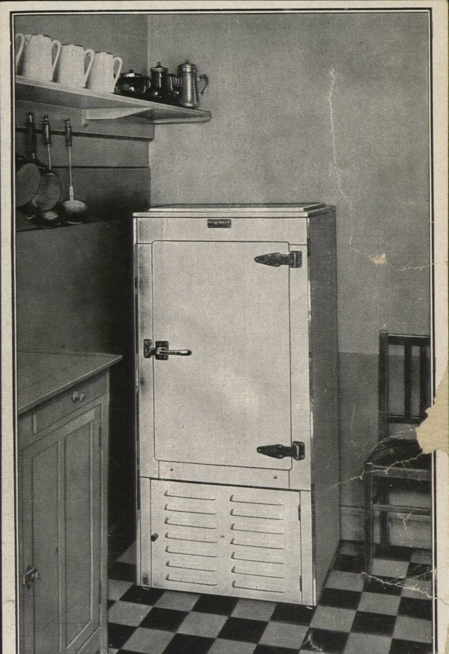 Black and white photo of an old-fashioned kitchen with a vintage refrigerator, a small wooden chair, shelves with cups and teapots, and checkered tile flooring.
