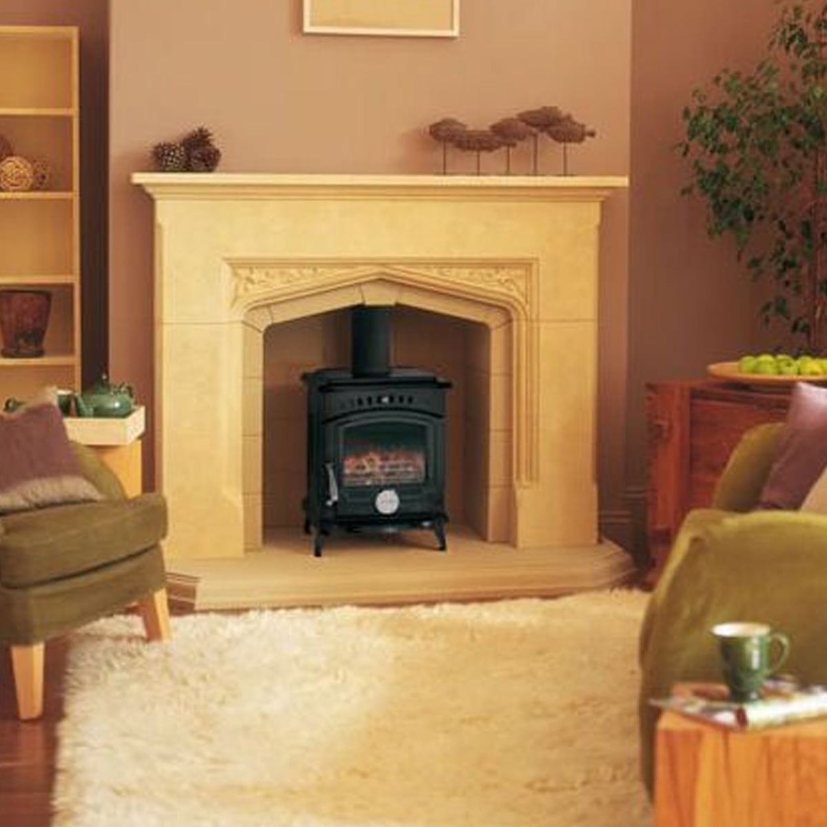 A cozy living room with a cream-colored fireplace, a black wood-burning stove, green armchairs, a white fluffy rug, and decorative items on shelves and the mantel. A potted plant sits in the corner.