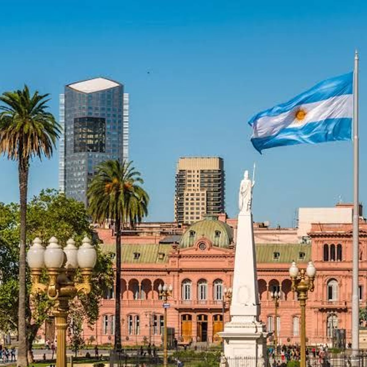 The Argentine flag waves near the Casa Rosada, a pink government building in Buenos Aires, with modern skyscrapers, palm trees, and a white monument in the foreground under a clear blue sky.