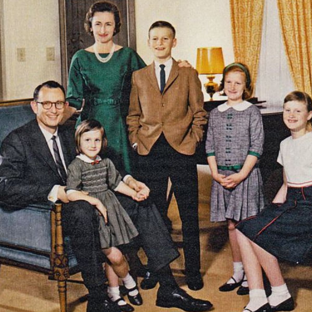 A family of six poses indoors; two adults stand and sit with four children in semi-formal 1960s-style clothing, smiling. The setting includes a lamp, curtains, and furniture in a warmly lit living room.