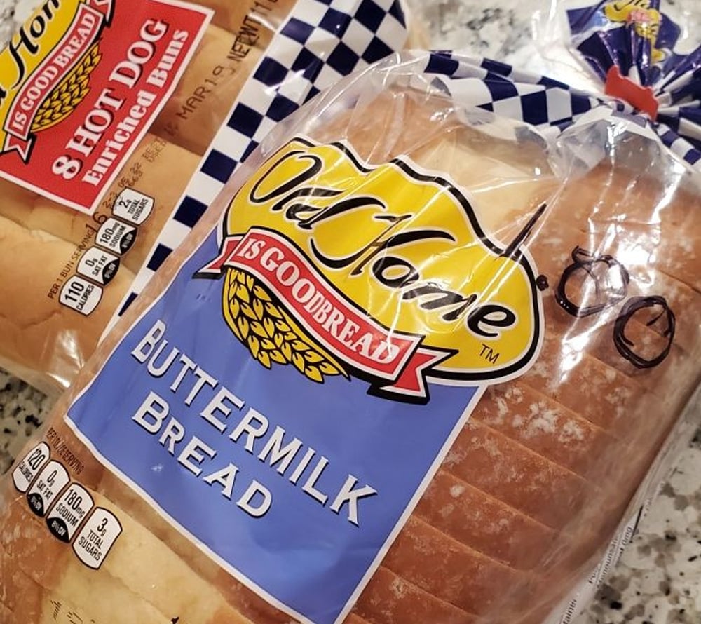 A loaf of Old Home Buttermilk Bread in clear packaging sits next to a package of hot dog buns on a countertop. The bread bag has a price written on it in black marker.
