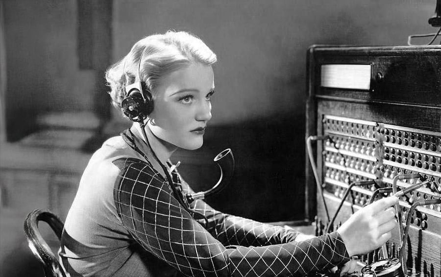 A woman wearing a headset operates a vintage telephone switchboard, connecting calls by inserting cables into jacks. She looks focused, with her hand reaching toward the switchboard, in a black-and-white setting.