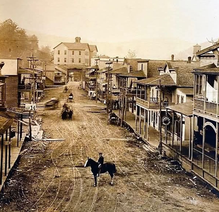 Sepia-toned photo of an old western town with dirt roads, wooden buildings, horse-drawn carriages, and a person on horseback in the street; hills and a large building are visible in the background.