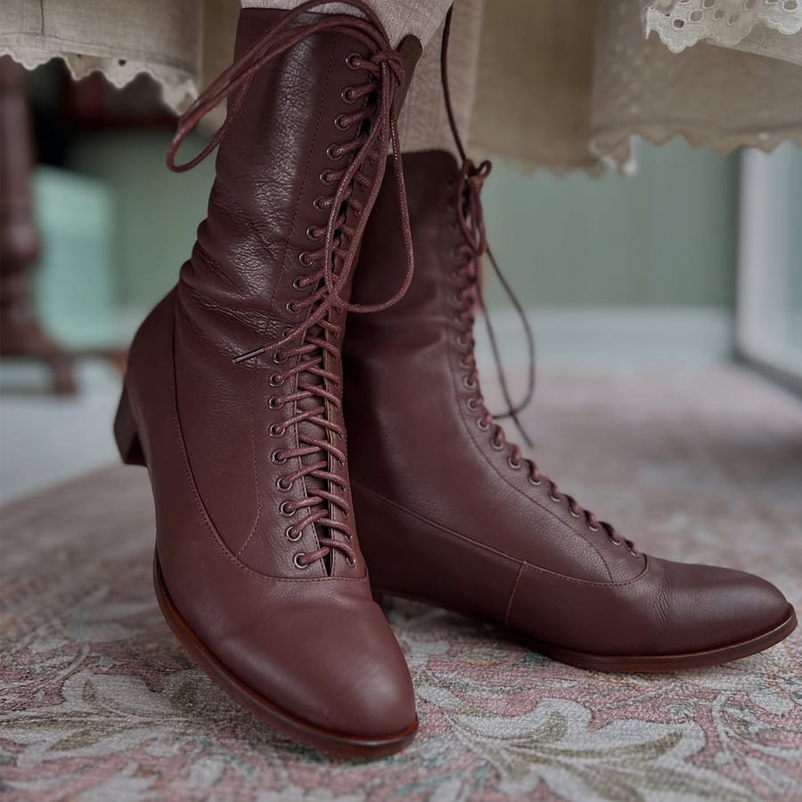 Close-up of a pair of vintage-style, brown leather lace-up ankle boots worn with a beige skirt with lace trim, on a patterned rug.