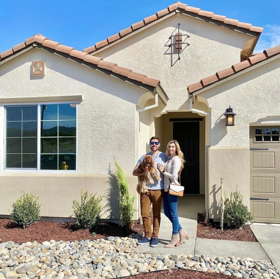 A smiling couple stands in front of a modern beige house, holding a brown dog. The sun is shining, and there are small plants and rocks in the landscaped front yard.