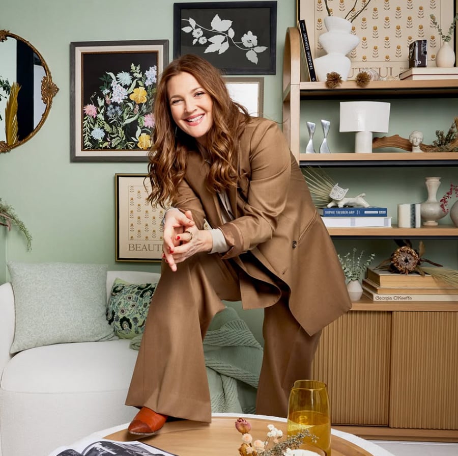 A woman in a brown suit smiles and leans forward in a cozy, stylish living room with botanical art, shelves of books, and decorative objects in the background.