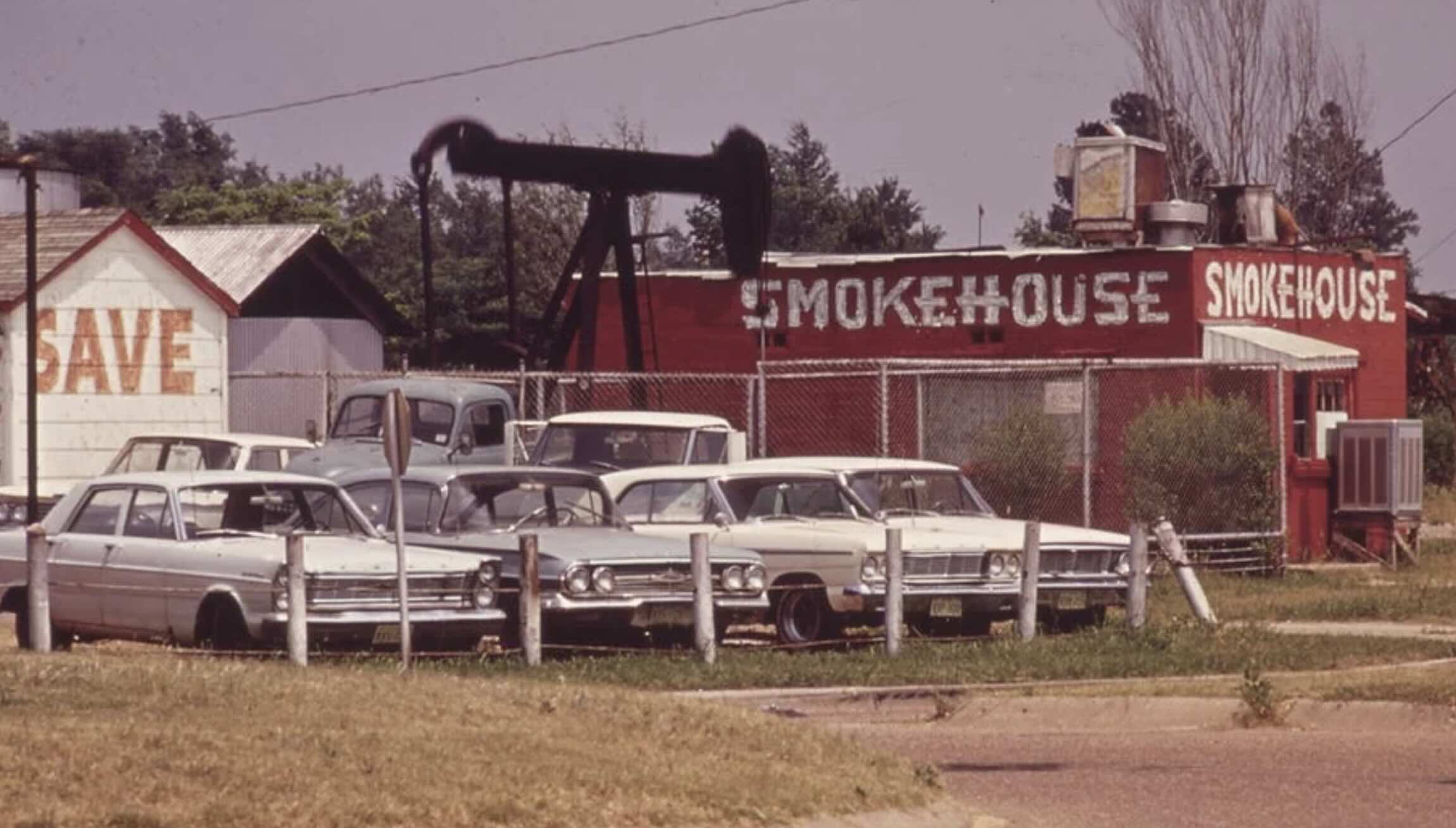 Several vintage cars are parked by a fence near an old smokehouse building with a sign. An oil pumpjack and a small "SAVE" painted structure are in the background, with trees and grass surrounding the scene.