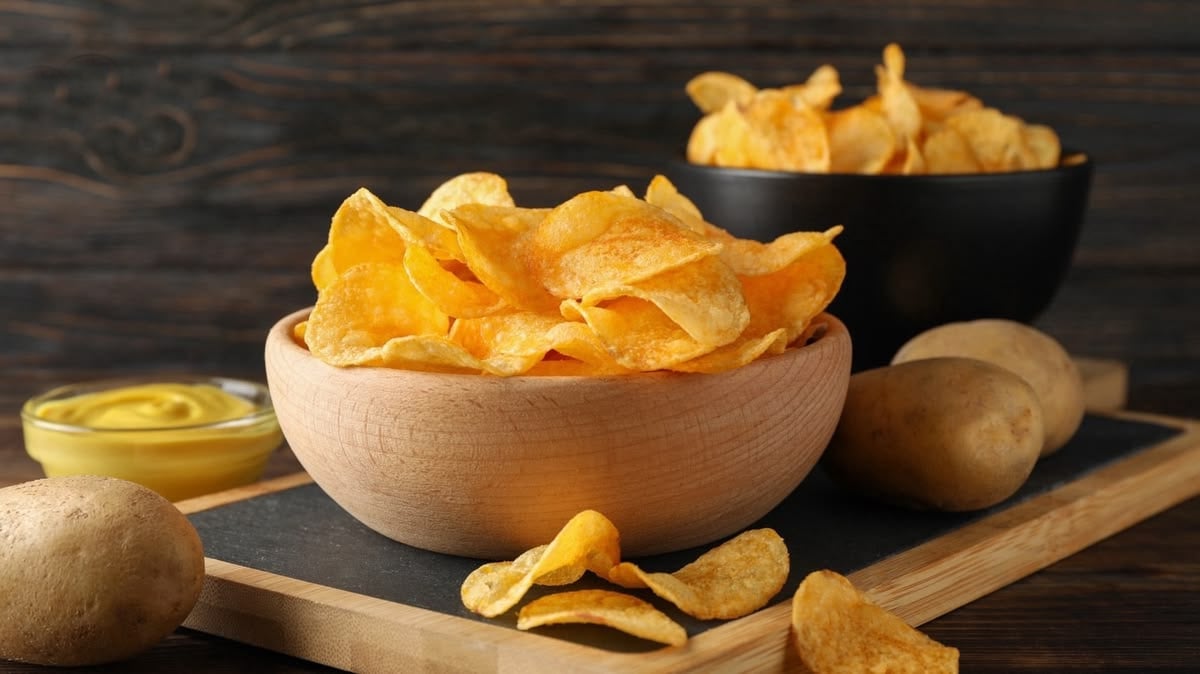 A wooden bowl filled with crispy potato chips sits on a slate board next to whole potatoes, a few loose chips, and a small dish of yellow dipping sauce, with another bowl of chips in the background.