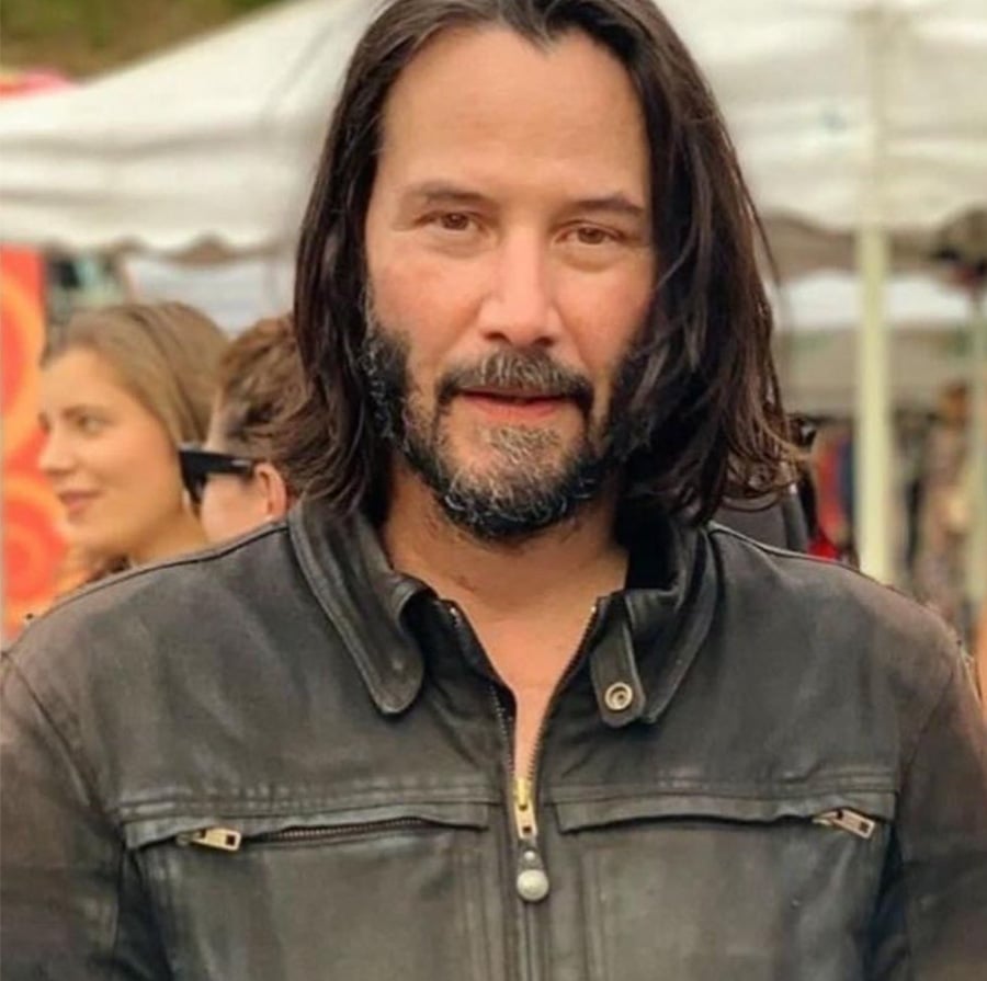 A man with shoulder-length dark hair, a beard, and a mustache is wearing a black leather jacket and standing outdoors, with people and tents blurred in the background.