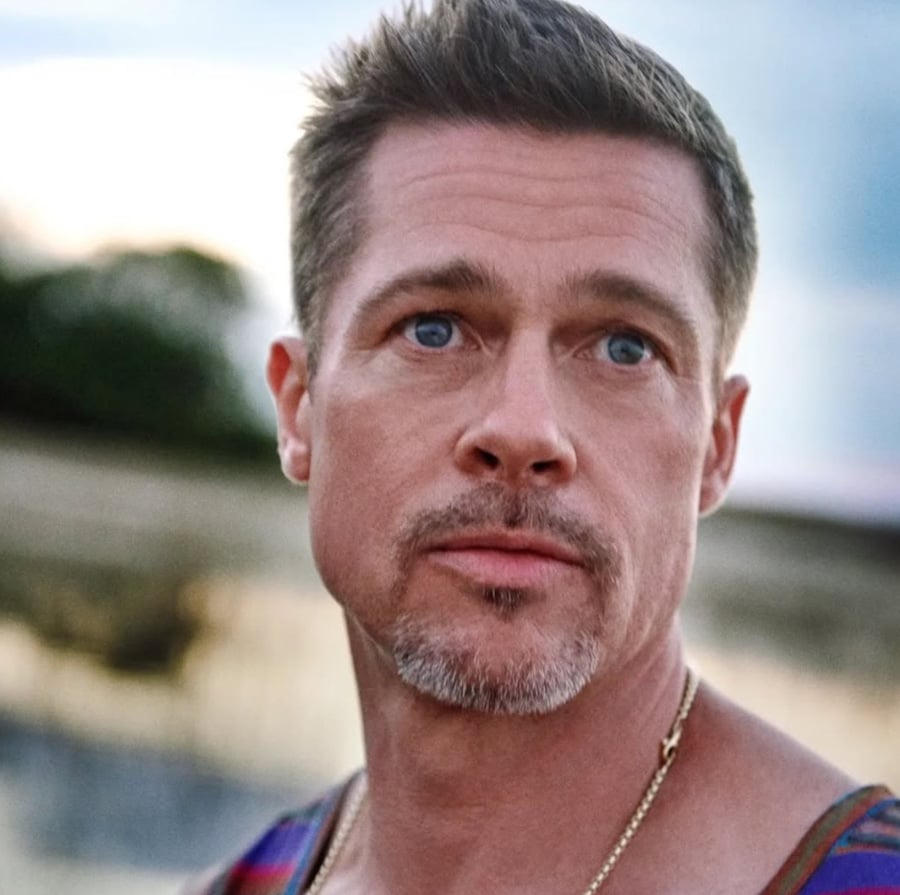A man with short brown hair, blue eyes, and light facial hair gazes slightly to the side outdoors. He wears a sleeveless shirt and a gold chain necklace, with a blurred natural background behind him.