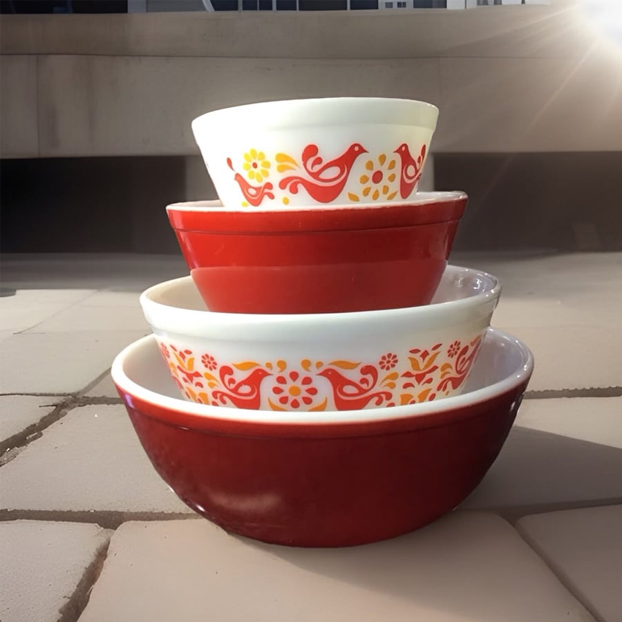 Four vintage bowls are stacked outdoors. The top and third bowls are white with red and yellow bird and flower patterns, while the second and bottom bowls are solid red. Sunlight highlights the bowls.