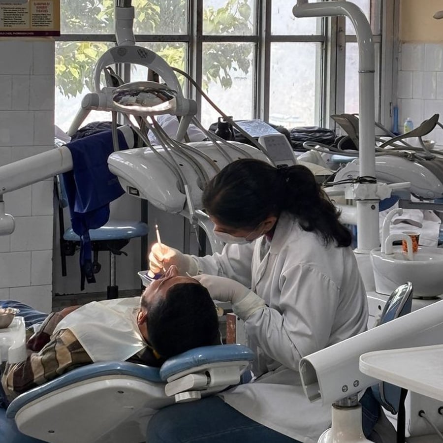 A dentist in a white coat examines a patient’s mouth in a dental clinic, using dental tools. The patient is seated in a reclined chair, and dental equipment surrounds them. Sunlight streams through the window in the background.