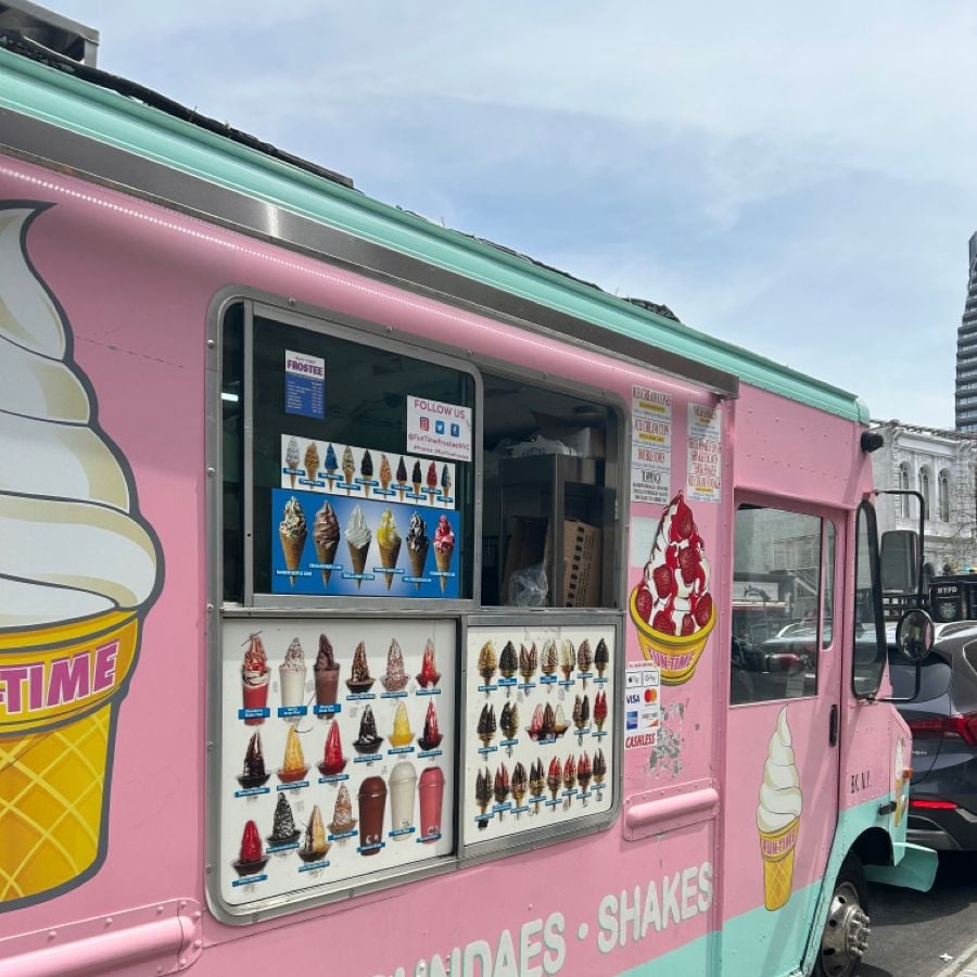 A pink and blue ice cream truck with a menu of various ice cream cones, sundaes, and shakes displayed on the side, parked on a city street. Part of a large ice cream cone graphic is visible on the truck.