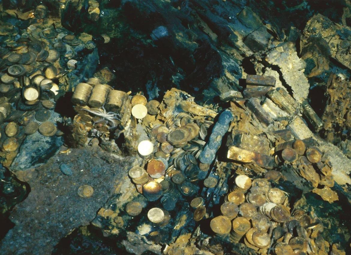 A collection of gold coins and bars scattered and stacked among rusted metal debris underwater, likely from a sunken shipwreck.